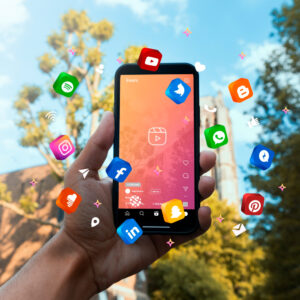 Foto van een hand met smartphone met eromheen plaatjes van logo's van verschillende apps en op achtergrond is de blauwe lucht en wat bomen te zien