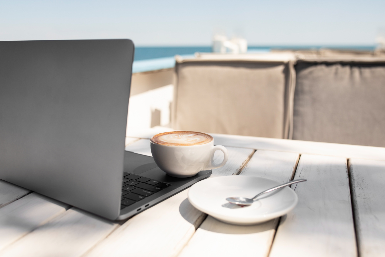 Een geopende laptop op tafel met een kopje koffie ernaast en op achtergrond het strand